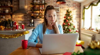 Eine junge Frau sitzt in weihnachtlich dekorierter Küche vor einem Laptop und wirkt konzentriert. Im Hintergrund steht ein geschmückter Weihnachtsbaum, Lichterketten und Kerzen sorgen für gemütliche Stimmung. Auf dem Tisch steht eine rote Tasse. Das Bild symbolisiert kreatives Arbeiten in der Adventszeit und passt zum Thema KI Prompts zu Weihnachten.