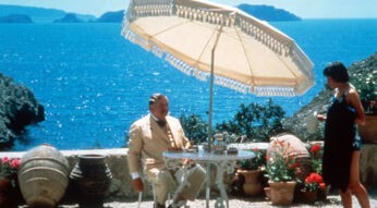 Filmszene aus "Das Böse unter der Sonne". Peter Ustinov, als Meisterdetektiv Hercule Poirot, sitzt auf einer sonnigen Terrasse an einem Tisch. Im Hintergrund ist das Meer zu sehen.