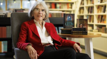 Donna Leon sitzt in einem Sessel und lächelt freundlich in die Kamera. Daneben steht ein kleiner Tisch mit einigen ihrer Bücher darauf.
