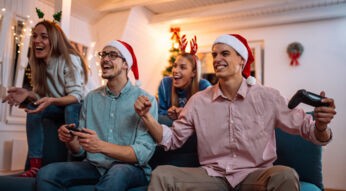 Auf dem Bild in dem Artikel zu den Spielen zu Weihnachten sind vier junge Erwachsene zu sehen, die auf einem Sofa sitzen und gemeinsam Videospiele spielen.