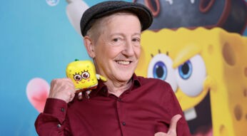 Auf dem Bild ist Santiago Ziesmer, der deutsche SpongeBob-Synchronsprecher, mit einem breiten Lächeln zu sehen. Er trägt ein weinrotes Hemd und eine Schiebermütze und hält ein kleines Stofftier von SpongeBob in der Hand. Im Hintergrund ist eine bunte Wand mit einem großen SpongeBob-Gesicht zu erkennen. Die Stimmung ist fröhlich und verspielt.