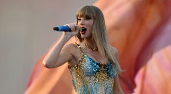 Das Bild zeigt Taylor Swift bei einem Live-Auftritt und dient symbolisch für Artikel über Taylor Swift-Dokus. Sie trägt ein glitzerndes, gold-blaues Bühnenoutfit und singt mit ausdrucksstarkem Gesichtsausdruck in ein blaues Mikrofon. Der verschwommene Hintergrund unterstreicht die emotionale und kraftvolle Atmosphäre der Szene.