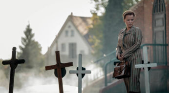 Die Bleikinder bei Netflix: Eine besorgte Frau steht mit einer Handtasche in den Händen an Kindergräbern vor einer Kirche.