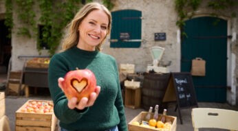 Hauptdarstellerin Diana Staehly steht auf dem Hof und hält lächelnd einen Apfel mit ausgestanztem Herz in der Hand.