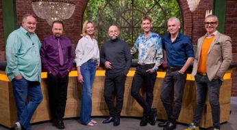 Auf dem Bild sind die Bares für Rares Händler Walter Lehnertz, Wolfgang Pauritsch, Susanne Steiger, Fabian Kahl, Christian Velten und Anaisio Guedes gemeinsam mit Moderator Horst Lichter zu sehen. Sie stehen gut gelaunt im Studio der ZDF-Sendung, vor einer Kulisse mit rustikaler Backsteinwand und großen Fenstern mit Blick ins Grüne.