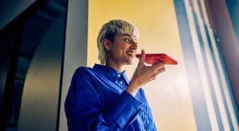 Eine lächelnde Person mit blondem Kurzhaarschnitt trägt ein leuchtend blaues Hemd und spricht in ein rotes Smartphone, als würde sie einen Sprachbefehl geben. Die Szene spielt sich in einem modernen Gebäude mit gelbem Hintergrund ab. Das Bild symbolisiert moderne Kommunikation und passt zum Thema Beste Sprachassistenten.