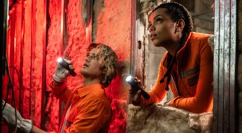Auf dem Bild zur Cold Storage Filmkritik sind Naomi Williams, gespielt von Georgina Campbell, und Travis „Teacake“ Meacham, dargestellt von Joe Keery, in einer spannungsgeladenen Filmszene zu sehen. Beide tragen orange Arbeitskleidung und leuchten mit Taschenlampen in einen dunklen, industriellen Raum. Die rote Beleuchtung und ihre konzentrierten Blicke erzeugen eine bedrohliche, nervöse Atmosphäre.