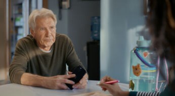Harrison Ford sitzt an einem Tisch. Er ist frontal zu sehen und hält ein Smartphone in der Hand. Er spricht mit einer nicht erkennbaren Person, deren Hand einen roten Stift hält.