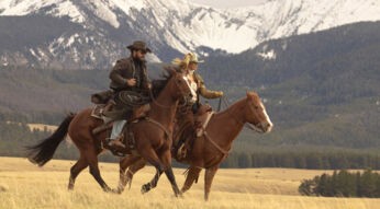 Auf dem Bild zur Dutton Ranch reiten Kelly Reilly als Beth Dutton und Cole Hauser als Rip Wheeler gemeinsam durch eine weite Landschaft. Beide sitzen auf Pferden vor einer beeindruckenden Bergkulisse mit schneebedeckten Gipfeln. Beth trägt einen Cowboyhut und wirkt fokussiert, während Rip entschlossen vorausblickt, was eine freiheitliche, zugleich kraftvolle Western-Stimmung vermittelt.