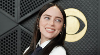 Auf diesem Bild ist Billie Eilish bei einem Event zu sehen. Sie trägt ein schwarz-weißes Outfit mit Hemd, Krawatte und Jacke und blickt mit einem freundlichen Lächeln leicht nach links. Hinter ihr befindet sich ein großes goldenes Grammophon-Symbol vor einer dunklen, strukturierten Wand. Das Bild wird symbolisch für den The Bell Jar-Film verwendet.