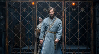 Auf dem Bild zu den GigaTV Highlights im Mai 2026 aus Der Medicus 2 ist Rob Cole, gespielt von Tom Payne, in einer düsteren, mittelalterlichen Szene zu sehen. Er steht im Vordergrund vor einem schweren Gittertor, trägt ein schlichtes graues Gewand mit Gürtel und wirkt erschöpft sowie nachdenklich. Im Hintergrund ist eine weitere Figur schemenhaft erkennbar, was die angespannte und geheimnisvolle Stimmung unterstreicht.