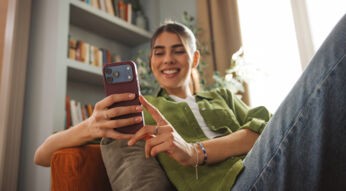 Auf diesem Bild zum Thema Handy auf Raten kaufen sitzt eine junge Frau entspannt auf einem Sofa in einem hellen Wohnzimmer. Sie hält ein Smartphone in der Hand und lächelt zufrieden auf den Bildschirm. Im Hintergrund sind ein Bücherregal und Pflanzen zu sehen, was eine gemütliche und positive Alltagsstimmung vermittelt.
