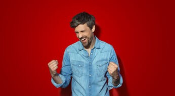 Auf dem Bild zum Angebot „Vodafone-Internet mit PS5 im Bundle“ ist ein Mann mit kurzen braunen Haaren und Bart zu sehen, der ein hellblaues Jeanshemd trägt. Vor einem roten Hintergrund ballt er begeistert die Fäuste und lacht.