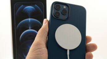 Auf diesem Bild zum Thema iPhone MagSafe ist ein blaues Apple iPhone in einer Hand zu sehen. Auf der Rückseite des Smartphones befindet sich ein weißes MagSafe-Ladegerät, das magnetisch befestigt ist und per Kabel verbunden wird. Im unscharfen Hintergrund steht die Verpackung des iPhones. Die Szene vermittelt einen modernen, technischen Eindruck und veranschaulicht die kabellose Ladefunktion des Apple MagSafe-Systems.