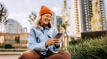 Auf dem Bild im Guide zum Thema unbegrenztes Datenvolumen sitzt eine junge Frau entspannt im Freien auf einer Grünfläche in einer modernen Stadt. Sie trägt eine orange Mütze, eine helle Jacke und Kopfhörer um den Hals, während sie lächelnd auf ihr Smartphone blickt.