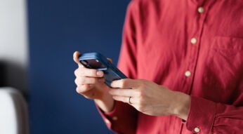 Auf dem Werbebild zu Apple Intelligence hält eine Person in einem roten Hemd ein blaues Smartphone in beiden Händen. Der Fokus liegt auf dem Gerät und der aktiven Nutzung, während der Hintergrund unscharf und ruhig wirkt. Die Szene vermittelt einen modernen, alltäglichen Technikmoment mit konzentrierter und persönlicher Stimmung.