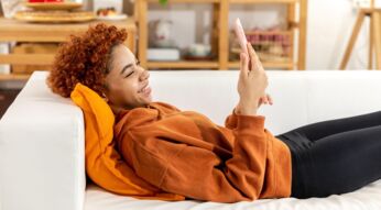 Schwarze Frau sitzt auf der Couch mit ihrem Smartphone und lächelt es an.