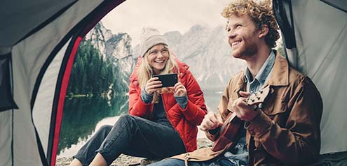 Junge Frau fotografiert Mann mit Gitarre im Zelt vor Bergsee mit Smartphone.
