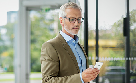 Business-Mann steht mit seinem Smartphone in der Hand am Firmeneingang und schreibt eine SMS.