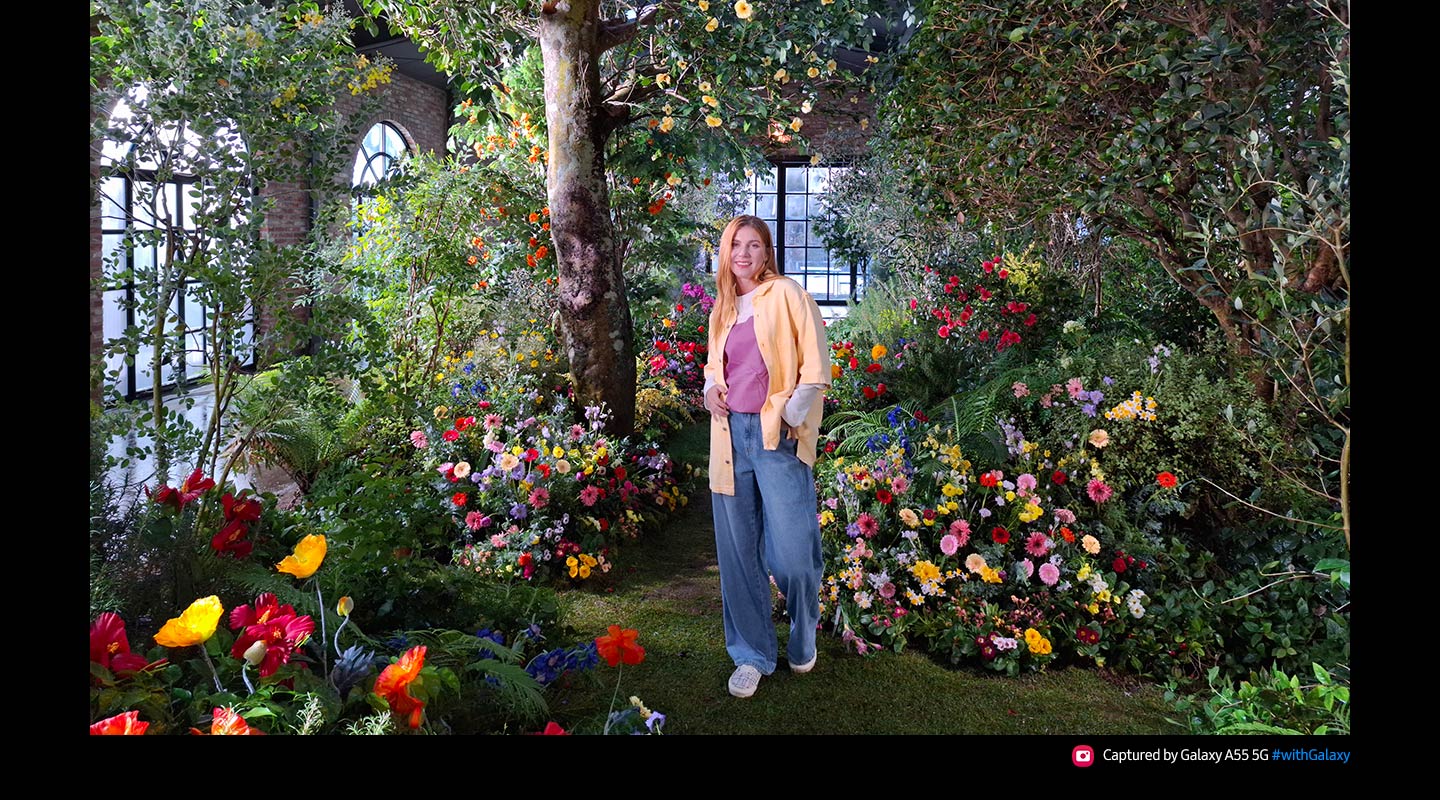 Ein mit 50 Megapixeln aufgenommenes, hochauflsendes Foto einer Person, die in einem ppigen Innengarten voller bunter Blumen und Grnem steht. Der Text liest: "Aufgenommen mit dem Galaxy A55 5G #withGalaxy".
