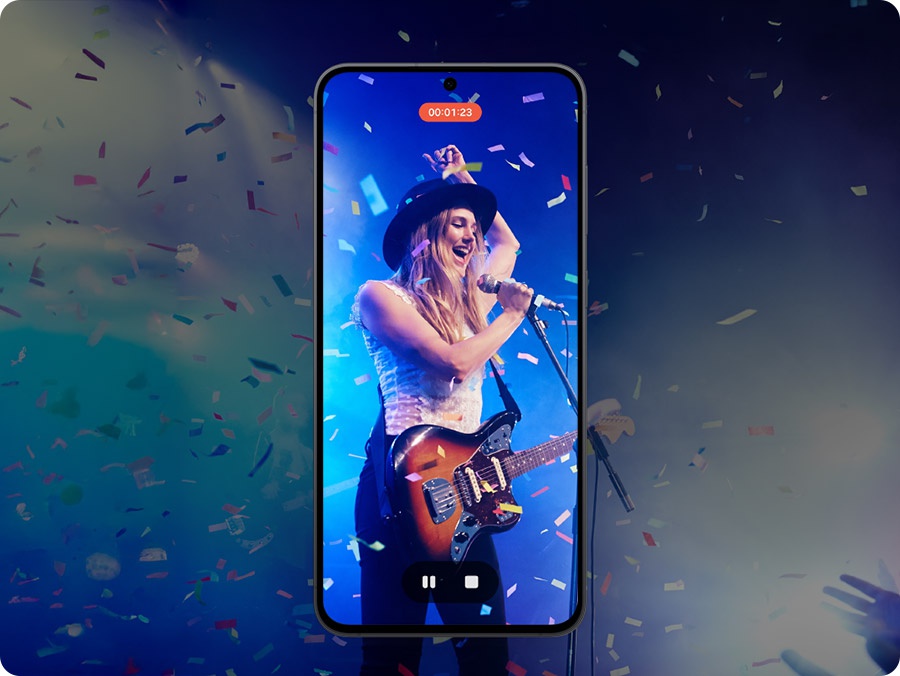 Auf dem Display eines Smartphones ist eine Videoaufzeichnung einer Live-Bhnenperformance zu sehen. Der*die Knstler*in trgt einen schwarzen Hut, spielt E-Gitarre und singt in ein Mikrofon. Blaue Bhnenlichter beleuchten den Hintergrund, whrend Konfetti herabfllt. Ein Timer zeigt 