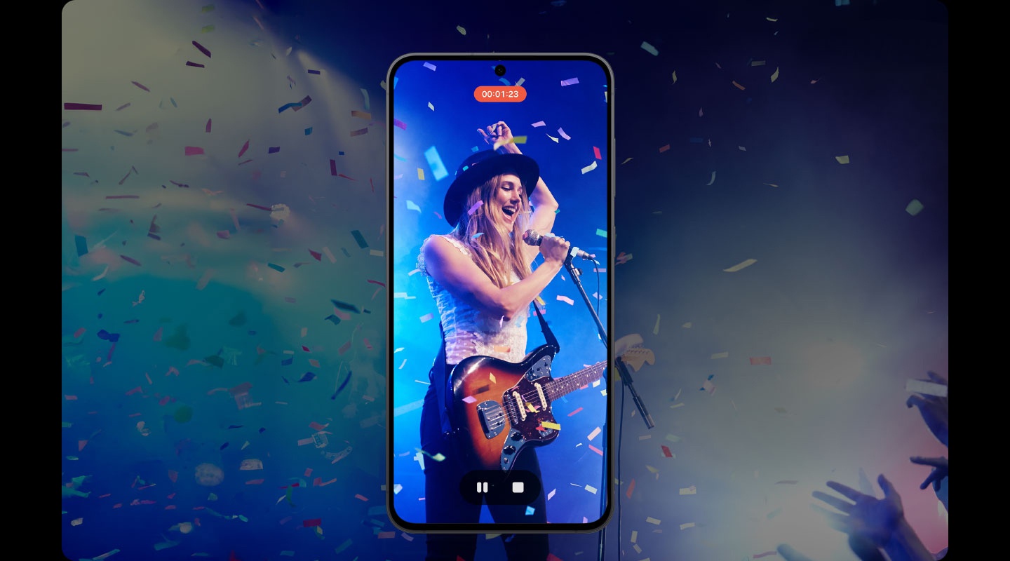Auf dem Display eines Smartphones ist eine Videoaufzeichnung einer Live-Bhnenperformance zu sehen. Der*die Knstler*in trgt einen schwarzen Hut, spielt E-Gitarre und singt in ein Mikrofon. Blaue Bhnenlichter beleuchten den Hintergrund, whrend Konfetti herabfllt. Ein Timer zeigt 