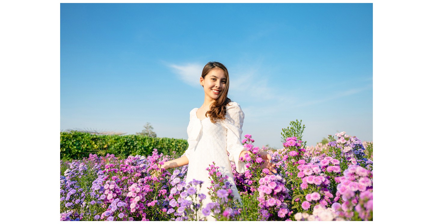 Eine Frau in einem wei�en Kleid steht drau�en zwischen vielen Blumen und posiert f�r ein Foto.

    