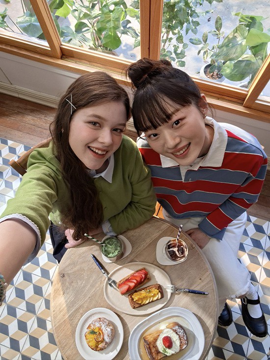 Selfie-Foto von zwei jungen Frauen beim Brunch in einem hellen Caf�, von oben aufgenommen mit der Frontkamera des Galaxy A57 5G. Die Bilddetails wirken lebendig und farbenreich dank Super HDR. Unten befindet sich ein Kamera-Icon mit dem Text 