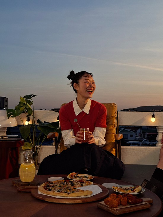 Foto einer l�chelnden jungen Frau auf einer Aussenterrasse bei wenig Licht, aufgenommen mit Galaxy A57 5G. Die Bilddetails sind hell und klar. Unten befindet sich ein Kamera-Icon mit dem Text 