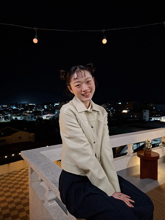 Foto einer jungen Frau auf einer Aussenterrasse bei Nacht, aufgenommen mit Galaxy A57 5G. Die Bilddetails sind dank optimierter Nightography hell und deutlich. Unten befindet sich ein Kamera-Icon mit dem Text 