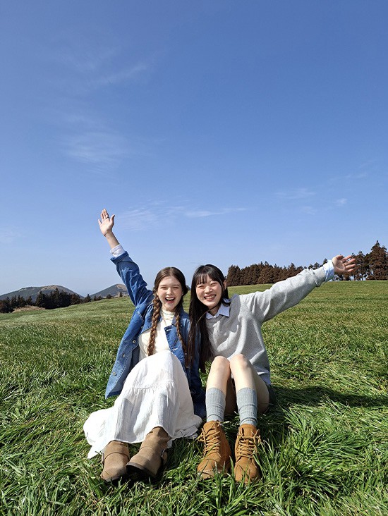 Foto von zwei jungen Frauen auf einer offenen Wiese vor blauem Himmel, aufgenommen mit der Weitwinkel-Kamera des Galaxy A57 5G. Die Bilddetails sind hell und lebendig mit verbesserter Qualit�t. Unten befindet sich ein Kamera-Icon mit dem Text 