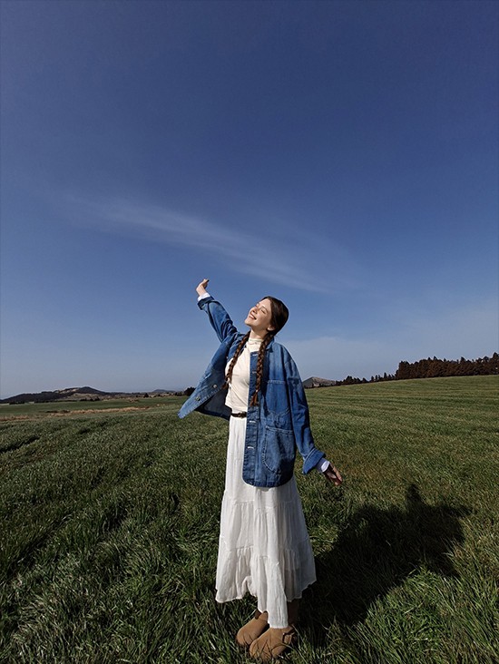 Foto einer jungen Frau auf einer offenen Wiese vor blauem Himmel, aufgenommen mit der Ultra-Weitwinkel-Kamera des Galaxy A57 5G. Die Bilddetails sind hell und lebendig mit verbesserter Qualit�t. Unten befindet sich ein Kamera-Icon mit dem Text 